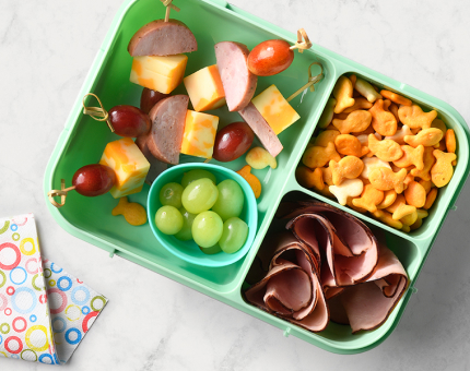 Green bento box filled with skewers of prepared sausage, cheese and grapes, mini cheese crackers and smoked ham slices.