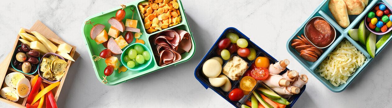 Four bento boxes on a white marble countertop including one wooden bento box filled with cheese, veggies, and hardboiled eggs, one blue box filled with grapes, humus and tomatoes and one green box filled with cold cuts and cheese skewers.