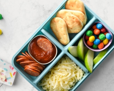 Light blue bento box filled with sauce, grated cheese, pepperoni, candies and mini naan breads on a white background.