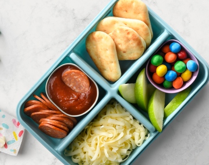 Light blue bento box filled with sauce, grated cheese, pepperoni, candies and mini naan breads on a white background.