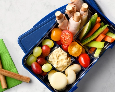Blue bento box with each compartment filled with breadsticks wrapped with turkey deli slices, cheeses, dip and veggies.