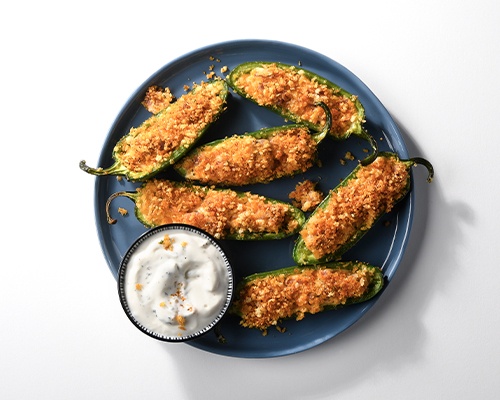 Blue plate with several crispy meat & cheese jalapeno poppers on top with a side ramekin of dipping sauce. 