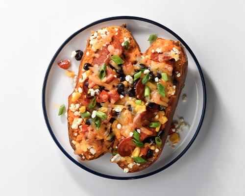 White, black rimmed plate with loaded baked sweet potatoes scattered with cheese, black olive pieces, green onions and more.