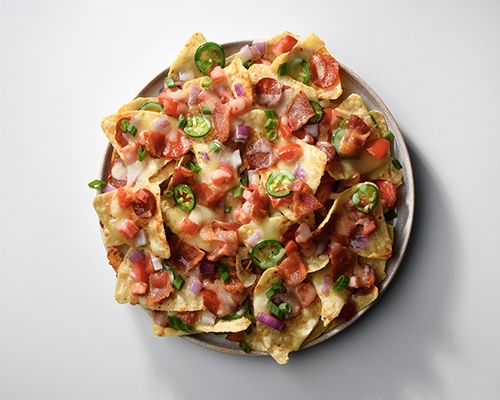 Gray and oat coloured plate full of nachos, melted cheese, sausage pieces, jalapeno pepper rounds and red onion.