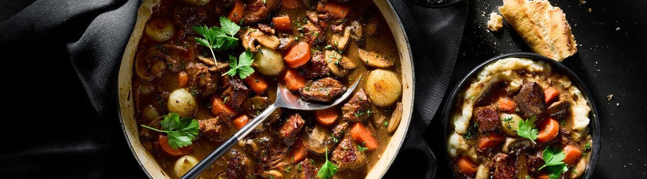 Black enamel dutch oven filled with Beef Bourgignon and a separate serving in a bowl where the beef tops a bowl of creamy, mashed potatoes.