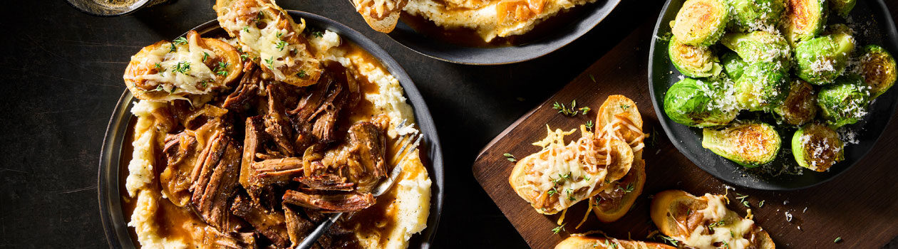 Two matte black bowls filled with creamy mashed potatoes and French Onion Pot Roast with sides of crostini topped with the caramelized onions from the roast, and a bowl of roasted Brussels sprouts.