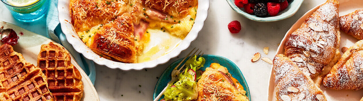 Three serving platters: one filled with waffle croissants, another with a croissant topped double cheese and ham strata and one with almond filled croissants.