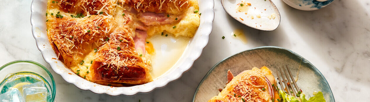 Ham and Cheese Croissant Strata in a white baking dish with a serving next to it on a gray dish with a side salad.