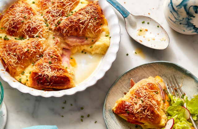 Ham and Cheese Croissant Strata in a white baking dish with a serving next to it on a gray dish with a side salad.
