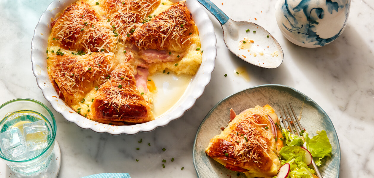 Ham and Cheese Croissant Strata in a white baking dish with a serving next to it on a gray dish with a side salad.