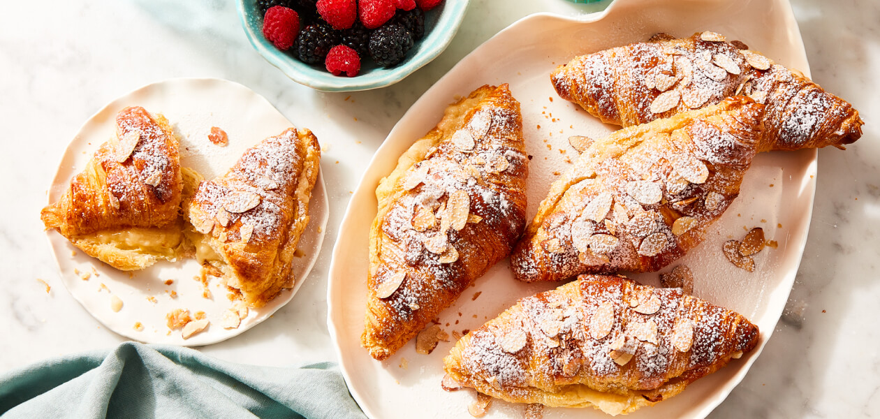 Speedy almond croissants topped with slivered almond slices in a cream serving dish next to a plate with one croissant on it.