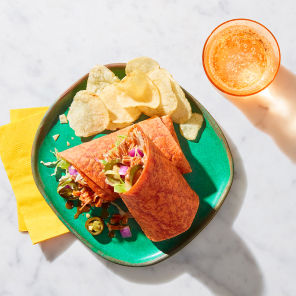 Red pepper wrap surrounding a maple bacon pulled pork filling with lettuce, red onions, cheddar and bbq sauce on a bright green plate next to a yellow napkin.