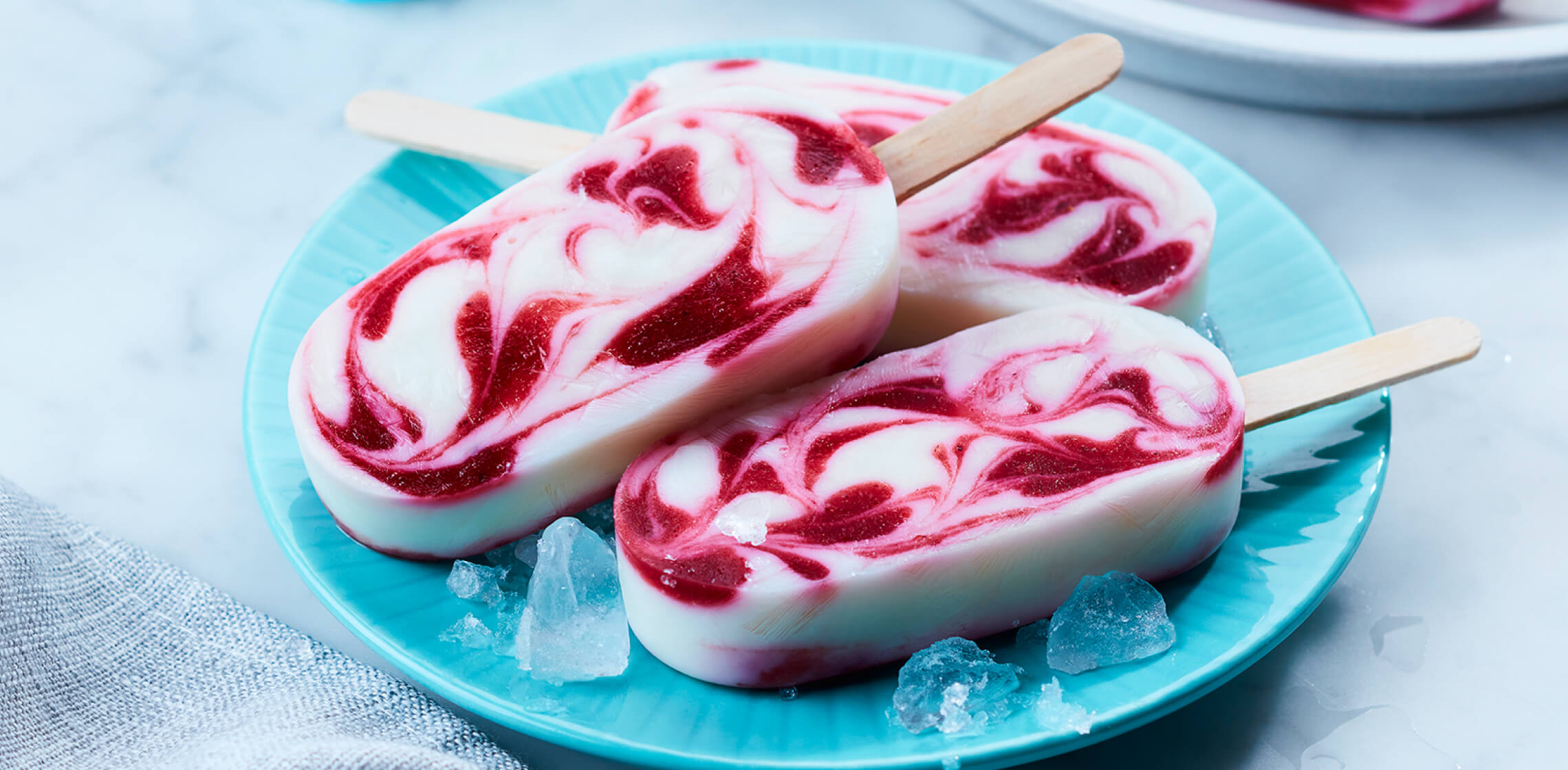 Swirled berry smoothie pops on a light blue plate with a few ice cubes around them.