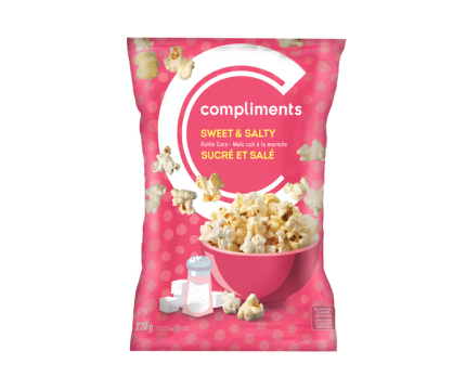 A pink packet of Compliments Sweet & Salty Kettle Corn, featuring an illustration of a salt shaker and sugar cubes on the front of the pack, along with a pink bowl of the popcorn.