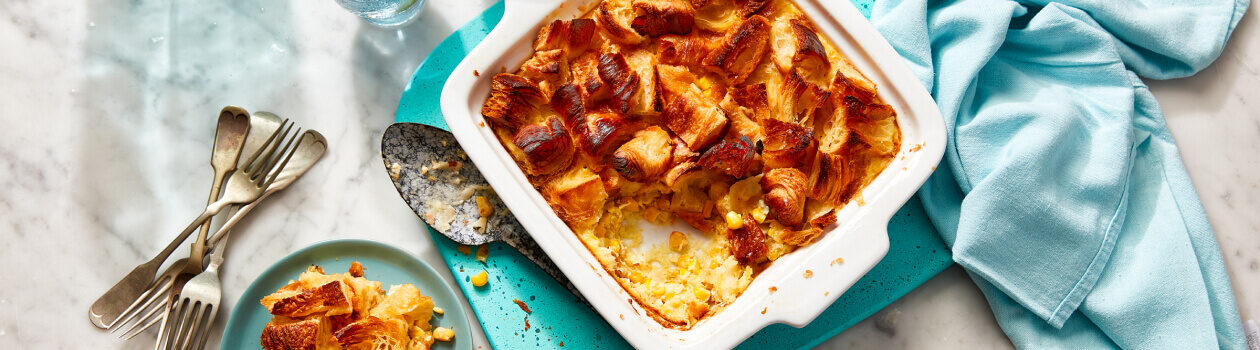 Croissant-topped corn pudding in a white baking dish with a serving next to it on a blue plate.