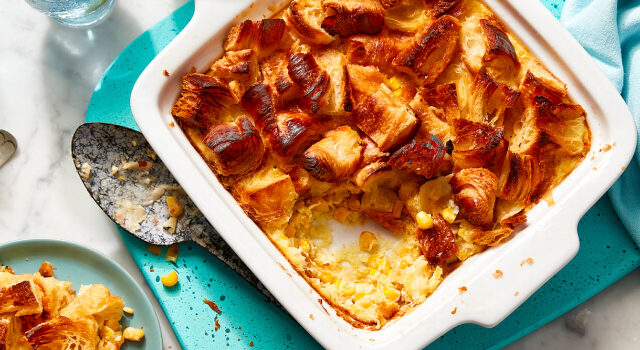 Croissant-topped corn pudding in a white baking dish with a serving next to it on a blue plate.