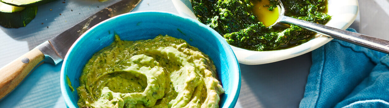 Two blue bowls of sauce including an avo tahini sauce and a fresh summer green herb sauce on a gray tray.