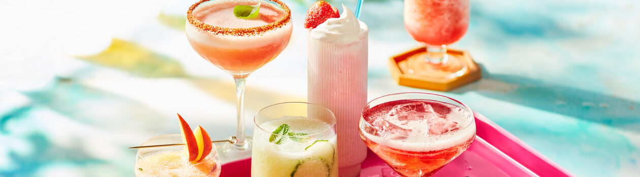 A selection of frosty summer cocktails in various glasses on a pink lacquered tray over a turquoise countertop.