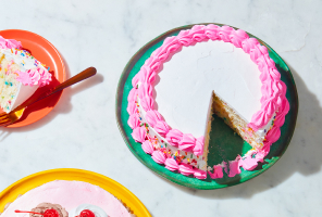 Triple-layer confetti cake iced in white and pink with sprinkles, sitting atop an orange dessert plate with a fork next to it.