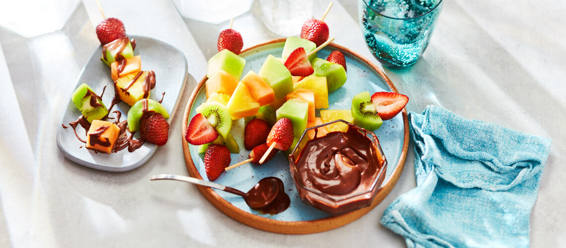 Various grilled fruit kabobs on a blue speckled plate next to a dipping bowl of chocolate sauce.