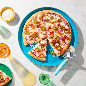 A family sized, maple bacon pulled pork pizza topped with tomato sauce, basil and pulled pork that sits atop a teal plate on a white marble countertop.