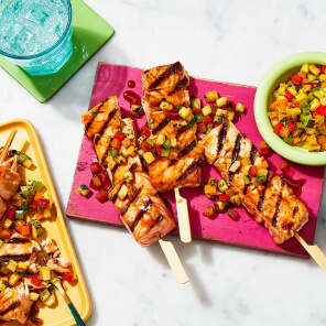 Pink board topped with teriyaki salmon skewers and a pineapple & spicy pepper kabob salsa.