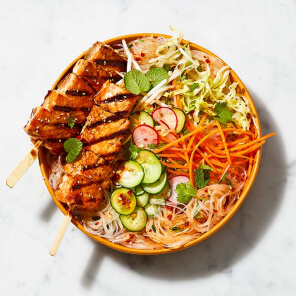 Orange plate topped with rice noodles and teriyaki salmon skewer, quick pickles and spiced salad dressing.