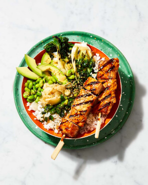 Orange bowl on top of a green plate filled with Teriyaki salmon skewers, short grain rice, baby bok choy, edamame and avocado.