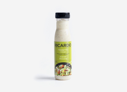 Clear glass bottle of Ricardo Caesar Salad Dressing with green label depicting a Caesar salad with croutons and Parm cheese shreds.