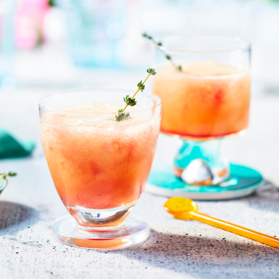 Glasses filled with a nectarine-hued Slushy Shirley cocktail and garnished with a sprig of thyme sitting on a grey countertop.