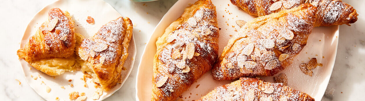 Speedy almond croissants topped with slivered almond slices in a cream serving dish next to a plate with one croissant on it.