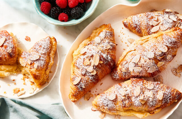 Speedy almond croissants topped with slivered almond slices in a cream serving dish next to a plate with one croissant on it.