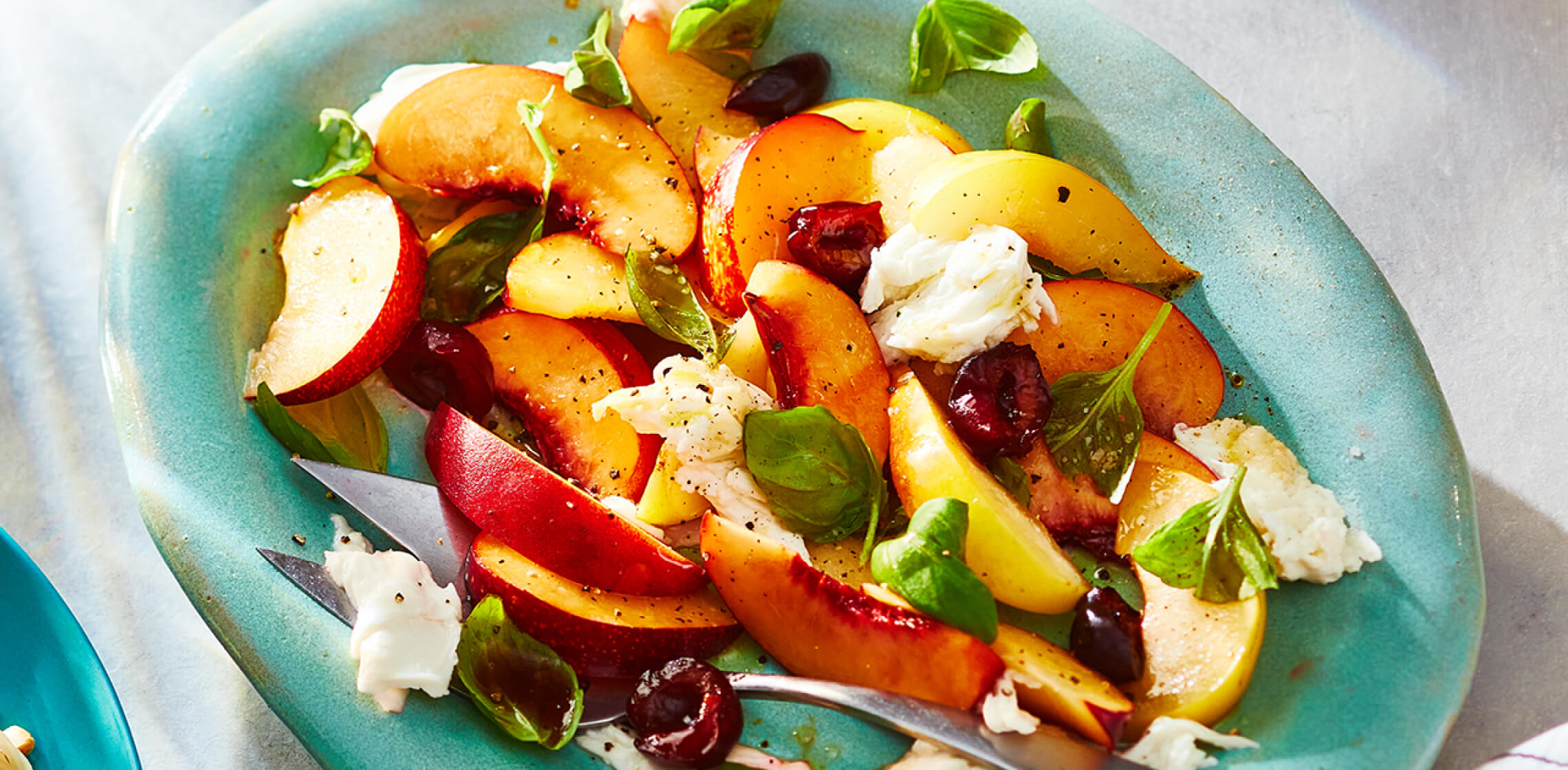 Light green serving platter topped with a fresh, stone fruit Caprese-style salad and serving spoons.