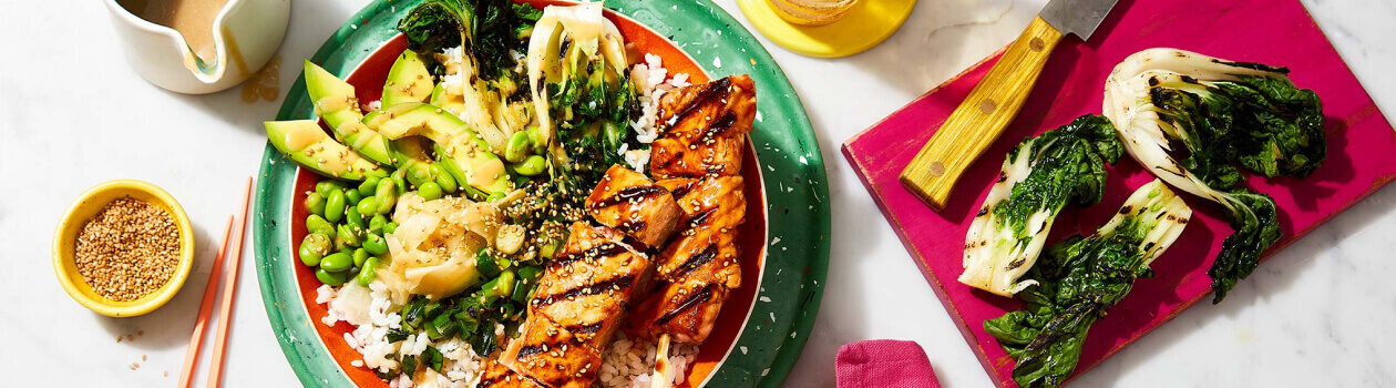 Orange bowl on top of a green plate filled with Teriyaki salmon skewers, short grain rice, baby bok choy, edamame and avocado.