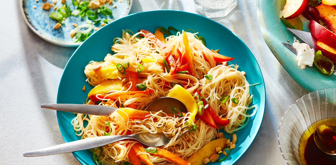 Blue plate topped with a Thai-inspired rice noodle salad sitting next to a plate with crushed peanuts and fresh herbs for toppings.