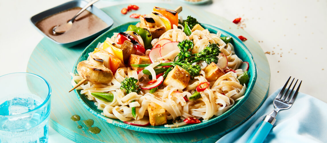 Fresh noodle bowl featuring grilled veggie and tofu skewers on top with bright broccoli florets on a turquoise plate.