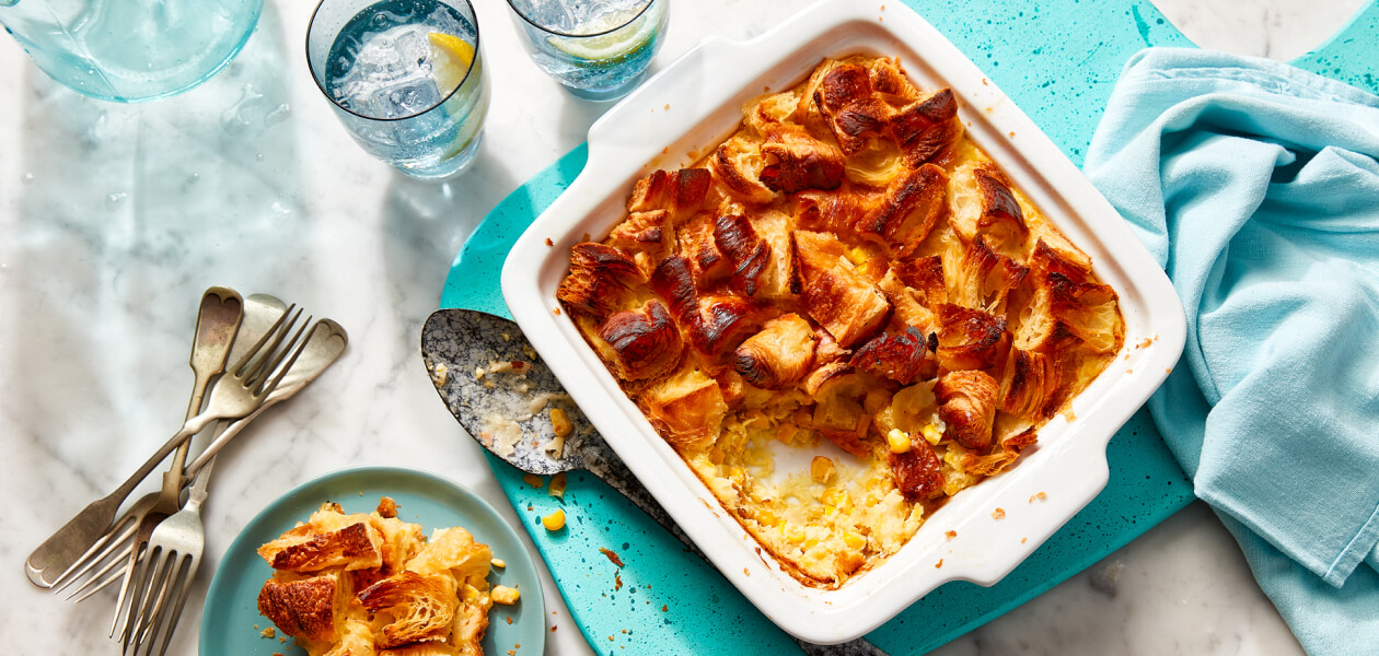 Croissant-topped corn pudding in a white baking dish with a serving next to it on a blue plate.