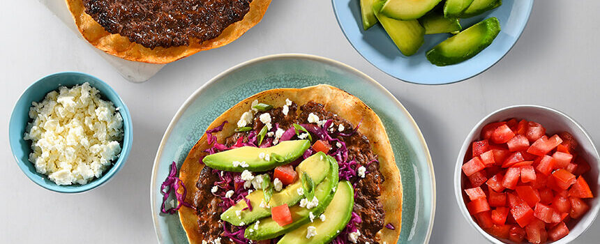 A photograph of a crisp flat taco topped with seasoned smashed beef, red cabbage, red onion, tomato, sliced avocado, and crumbled feta cheese for a colourful and delicious meal. Surrounding the plate is four smaller plates each containing feata cheese, chopped tomatoes, sliced avocados, and other loaded smash beef tacos as it