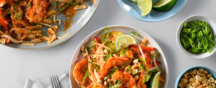 A colourful bowl of shrimp pad thai garnished with green onions, sliced lime, been sprouts, red peppers, and more. Around the place setting is more bowls of the individual garnishes to each dish can be to-taste!