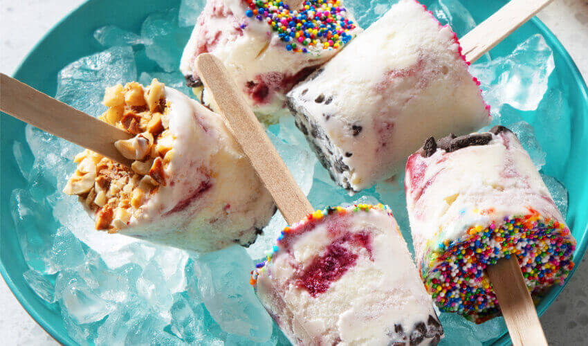 An assortment of frozen yogurt-based pops topped with sprinkles, chocolate bits, nuts and candy on ice in a blue bowl and one pop off to one side in an orange bowl.