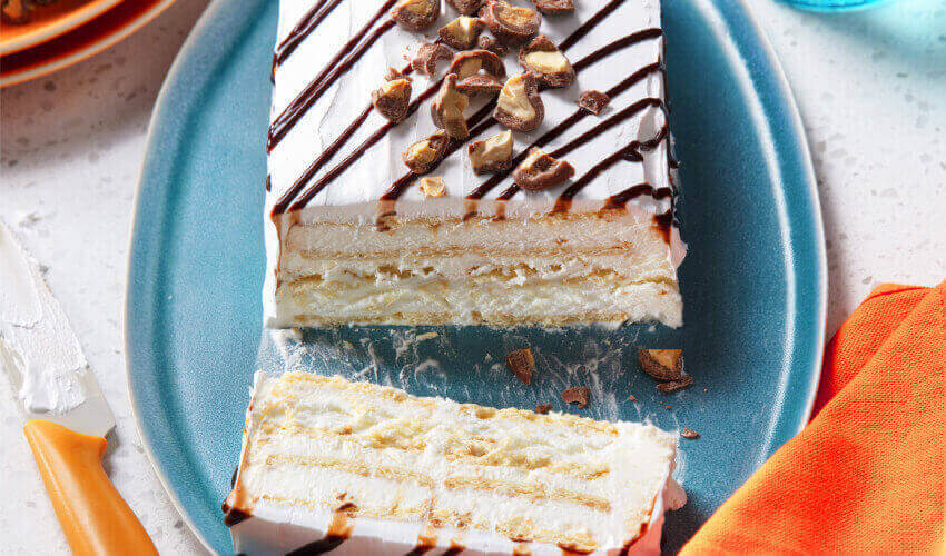 Choco-caramel layered ice cream cake using Compliments ice cream sandwiches, topped with a drizzle of sundae chocolate topping on a blue plate with the first slice cut.