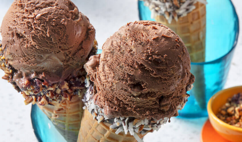 Dressed up waffle ice cream cones dipped in chocolate and covered in mixed nuts and coconut are topped with big scoops of chocolate ice cream, standing vertically in two blue glasses with a used ice cream scoop sitting next to it.