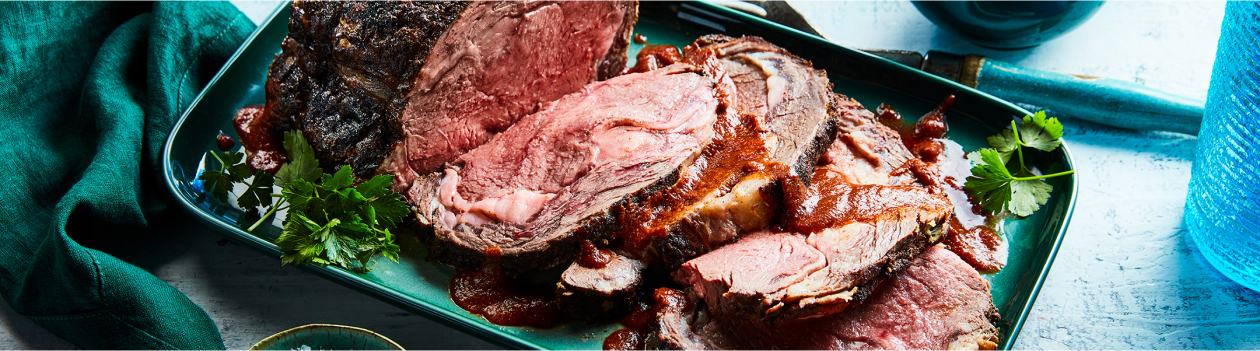 Rectangular aqua green serving platter with smoky Berbere spiced prime rib beef roast with slices cut from one end, sauce overtop and green herb garnish on the side