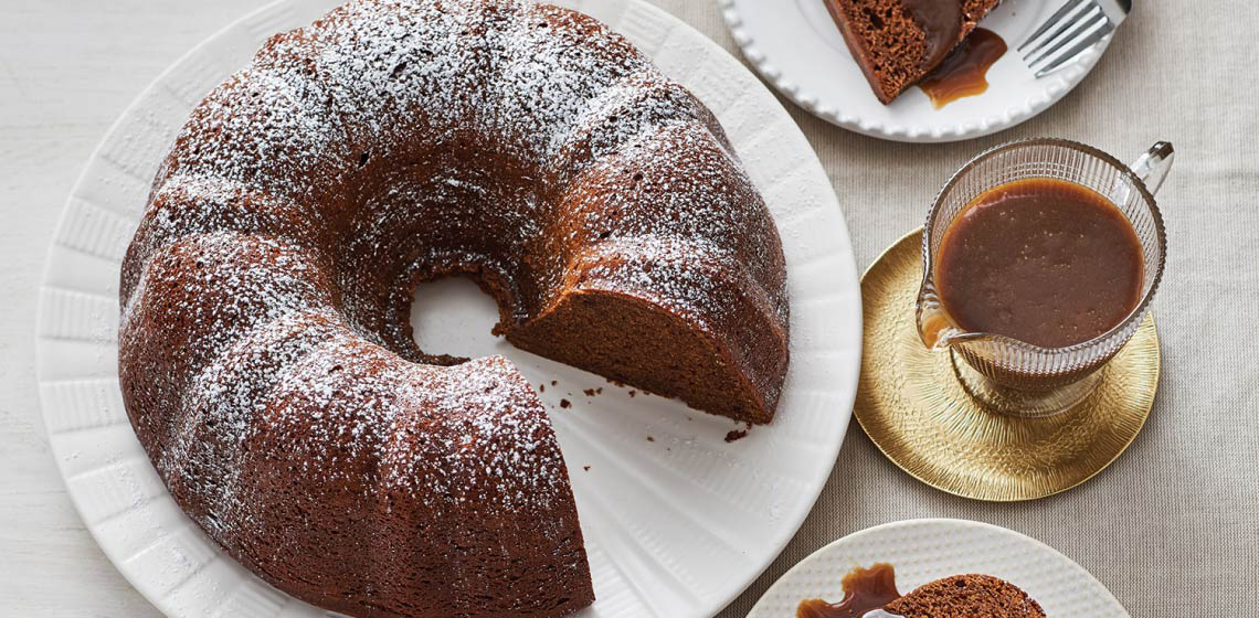 Gingerbread Bundt Cake sitting on a white cake plate, dusted with icing sugar and next to a container full of Coffee Caramel Sauce. 