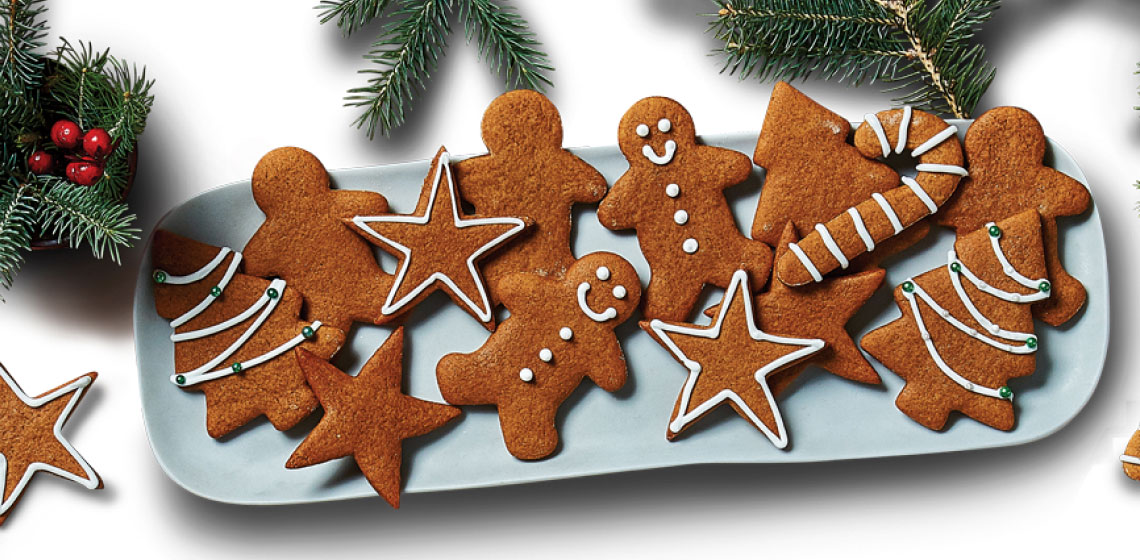 Gingerbread people, stars, and candy canes iced in white designs on an aged brown wooden plank on white plates.