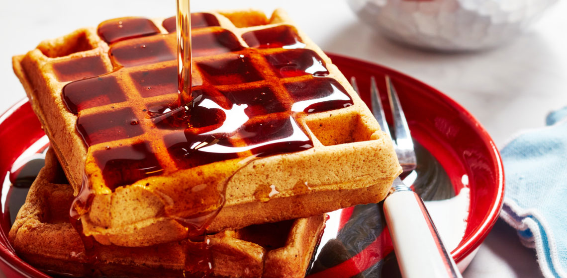 A close-up of a gingerbread waffle cut into by a fork on a white plate topped with maple syrup next to a small white bowl of fruit.