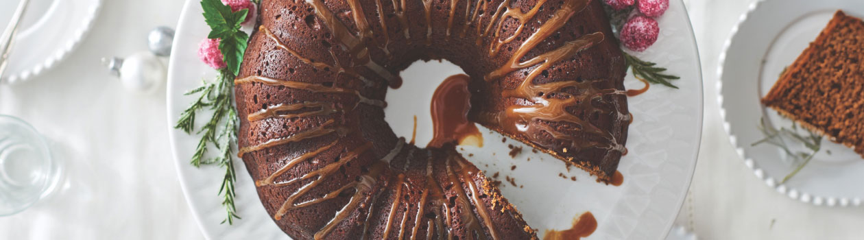 Gingerbread Bundt Cake sitting on a white cake plate, dusted with icing sugar and next to a container full of Coffee Caramel Sauce.