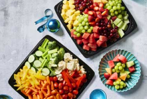 A vegetable and fruit tray side by side on a grey table beside plates and cups.