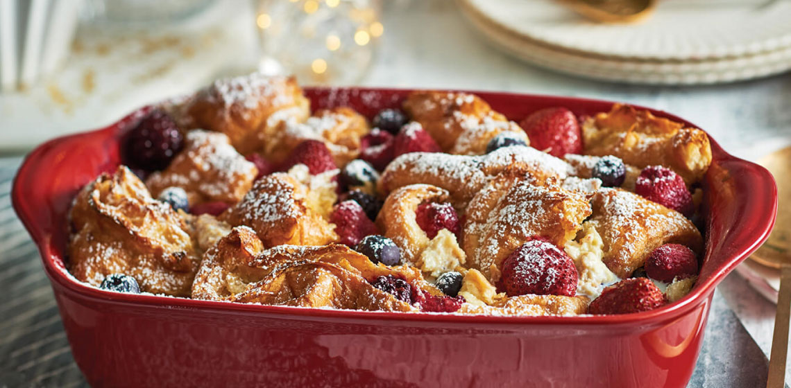 A red baking dish filled with a jumbleberry-dotted croissant strata dusted with icing sugar.