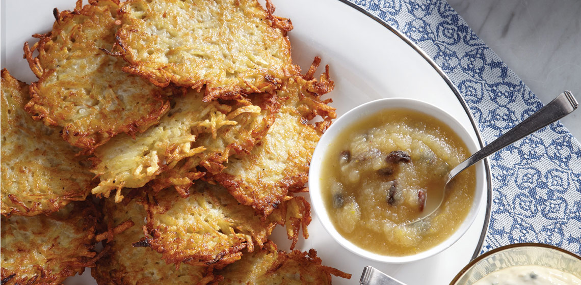 A white plate of freshly fried latkes next to a bowl of spiced applesauce with a spoon in it.